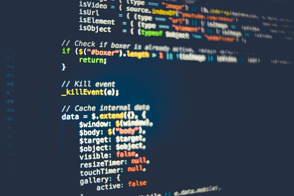 A close-up of a person's hand typing on a laptop keyboard placed on a wooden surface. The laptop screen displays lines of code with a dark background and multicolored text. The individual is wearing a