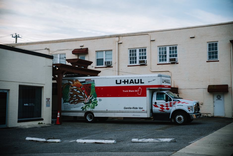 A white moving van with colorful graffiti on its side is parked on a paved street in front of a multi-storey red brick residential building. The van has an enclosed cargo area with no visible loading
