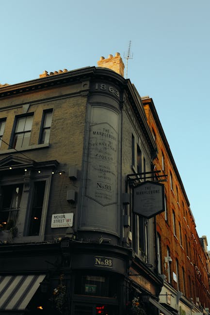 Photograph of a three-story building on a street corner, featuring traditional Georgian-style architecture with cream and brick exteriors. The corner façade displays a large, vintage-style sign indic